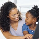 A mother and her young daughter both looking at each other smiling while on the computer.