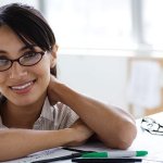 A woman wearing glasses, standing up leaning against a shelf with a pen and some paper placed in front of her.