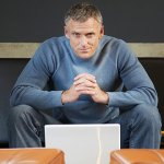 A man sitting down, working on his laptop looking into the camera.