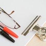 A white clipboard with glasses and a black and red pen sitting on top of it.