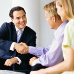 A meeting with a man wearing a business suit shaking hands with another man and a woman that are sitting together.