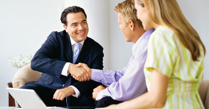 A meeting with a man wearing a business suit shaking hands with another man and a woman that are sitting together.