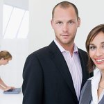 A man and a woman standing together in a white office space, with another woman in the background on her computer.