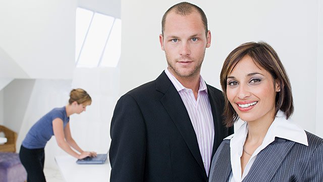 A man and a woman standing together in a white office space, with another woman in the background on her computer.