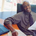 A man sitting in his office holding documents while smiling into the camera.