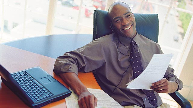 A man sitting in his office holding documents while smiling into the camera.