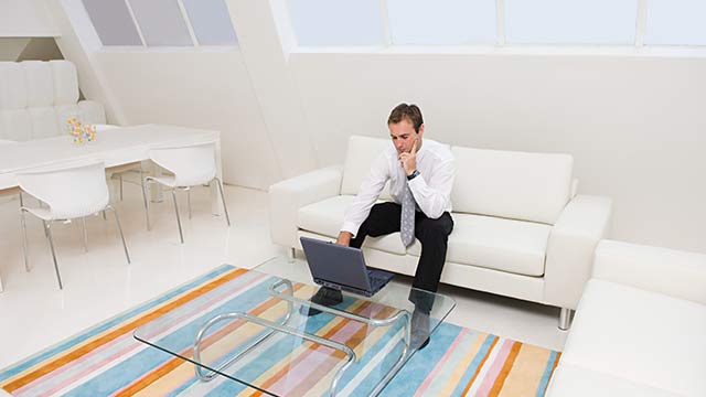 A man in a white room with a rainbow-colored carpet, sitting on a white couch using his laptop.