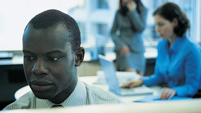 A man in an office working at a desk other employees are in the background working together.