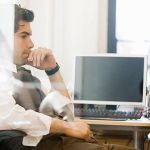 A man sitting in his office chair with a computer screen turned off behind him, thinking about the benefits of six sigma with his hand on his chin.