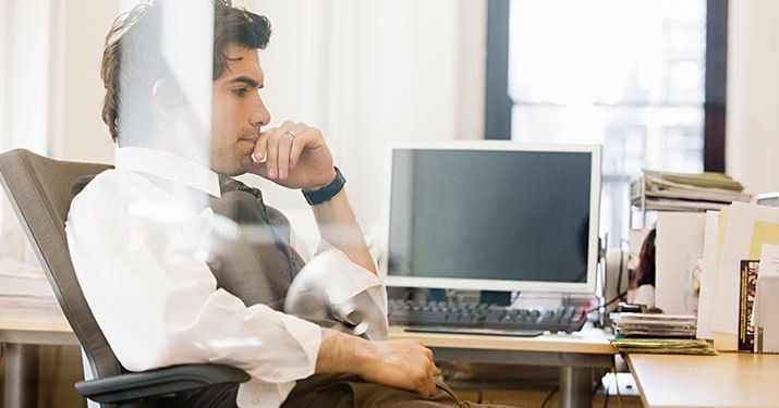 A man sitting in his office chair with a computer screen turned off behind him, thinking about the benefits of six sigma with his hand on his chin.