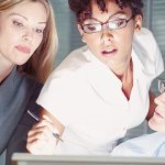 A man sitting at a computer with two women standing beside him helping each other figure something out on the computer screen.