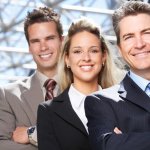 Three business people wearing suits, two men and one woman, smiling at the camera with a large artsy steel dome behind them.