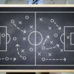 A person holding a small chalkboard with a soccer field drawing in chalk of x's and o's to represent a game plan.