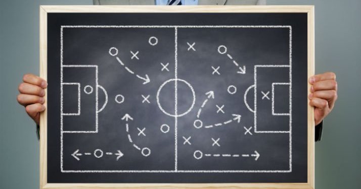 A person holding a small chalkboard with a soccer field drawing in chalk of x's and o's to represent a game plan.