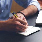 Close up of a man's hands writing on a piece of paper next to a laptop on a black desk.