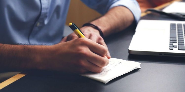 Close up of a man's hands writing on a piece of paper next to a laptop on a black desk.