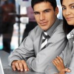 A man and woman in matching gray business clothes sharing a laptop and looking into the camera.