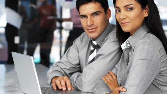 A man and woman in matching gray business clothes sharing a laptop and looking into the camera.