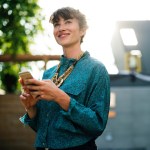 A woman outside on a college campus on her cellphone looking up on a sunny day smiling and laughing.