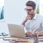 A man wearing glasses and a white button up shirt while working on his laptop at his desk.