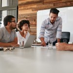 A man standing up in a boardroom with a whiteboard behind him giving a presentation about business process management to his colleagues.