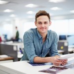 A man wearing a blue button up shirt smiling into the camera standing behind a desk in an open office space.