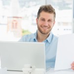 A man sitting at desk with a laptop holding a piece of paper smiling into the camera with an office window behind him overlooking a city.