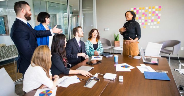 An agile project management meeting in a room with a large table and a group of employees on one side of the desk and one woman on the other side of the table giving a presentation -- behind her is a wall and a cluster of post it notes stuck to the wall.