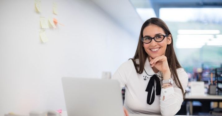 A scrummaster working on her computer with post it notes on the wall to help her plan the next agile sprint.