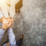 Overhead view of a group of people with their hands in the circle on top of each other as a representation of teamwork and globalization of HR leadership.