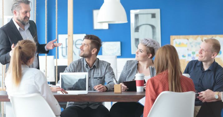 A modern workspace with vibrant blue walls and a group of co-workers sitting at a table looking up at a man standing up giving them instructions.