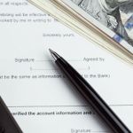 Close up of a contract with a pen sitting on top of the signature line, with a stack of hundred dollar bills and a calculator sitting on top of the contract.