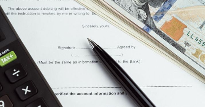 Close up of a contract with a pen sitting on top of the signature line, with a stack of hundred dollar bills and a calculator sitting on top of the contract.