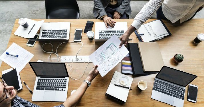 An overhead shot of a large cluttered desk with laptops, books, papers, phones, and coffee cups and two people handing paper to each other across the desk.