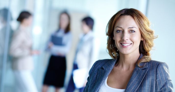 A business woman smiling into the camera with three other professionals out of focus in the background..