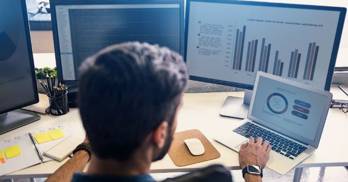 A man sitting at a desk with four computers evaluating various charts and data.