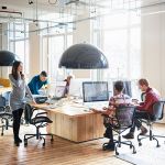 A modern office with large windows and five people sitting around a large desk with computers, working on managing virtual project teams.