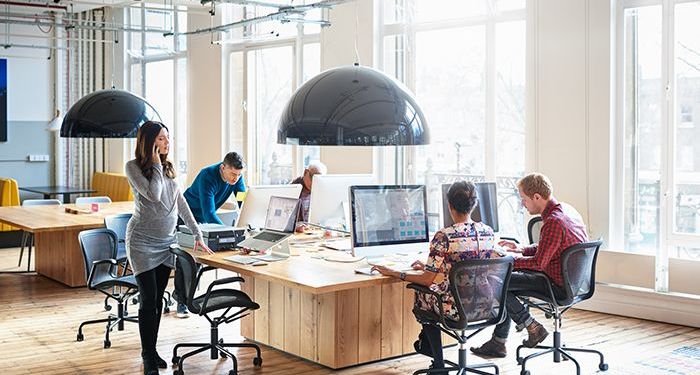 A modern office with large windows and five people sitting around a large desk with computers, working on managing virtual project teams.