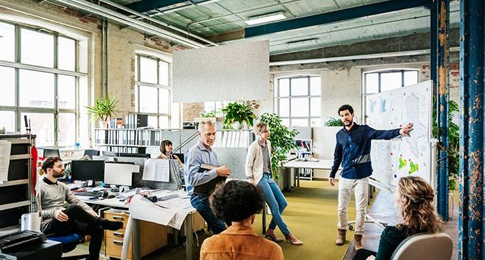 An urban office with messy desks and a group of employees sitting in office chairs gathered around a man giving a presentation in front of a board with information on it.