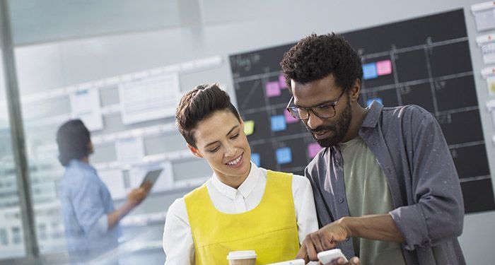 A woman wearing a yellow vest is holding a tablet and a cup of coffee while helping a project management coworker with scheduling on his smartphone.