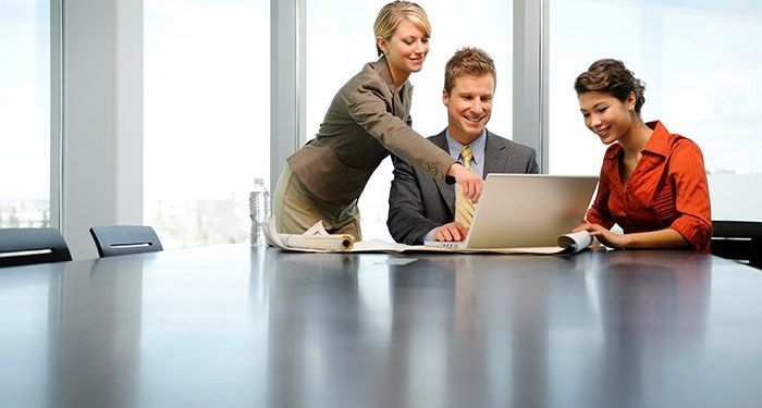 Three people in a highrise office at a board table all looking at a laptop with a window behind them overlooking a city.