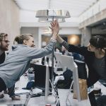 Project managers in an office space reaching across desks to congratulate each other with a high five.