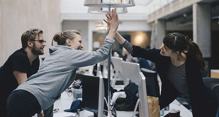 Project managers in an office space reaching across desks to congratulate each other with a high five.