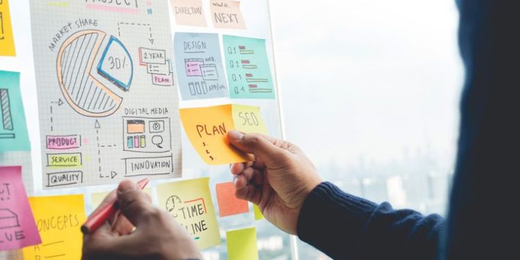 A project manager visualizes the iron triangle of project management by posting multi-colored sticky notes to a whiteboard.