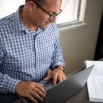 Adult learner at home reading textbook and using laptop.