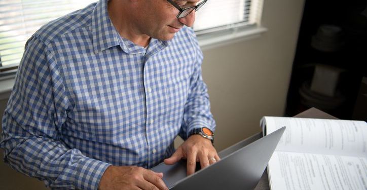 Adult learner at home reading textbook and using laptop.